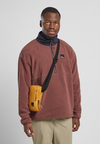 Bruin fleece trui met een marineblauwe kraag, ritssluiting en zwart logo. Gele tas bevestigd met een riem, beige broek, neutrale achtergrond.