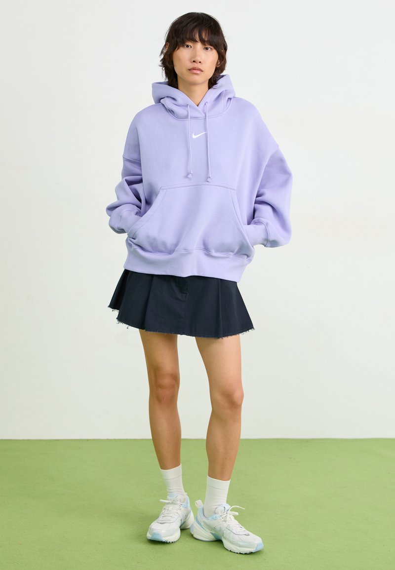 Sudadera de gran tamaño en color púrpura con bolsillo frontal y logo blanco, combinada con una falda negra de pliegues y calcetines blancos. El modelo está de pie sobre un suelo verde.