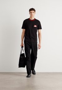 Black T-shirt with red collar and chest graphic, paired with loose black cargo pants and black shoes, carrying a black tote bag.