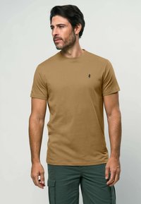 Krūškurvja T-krekls ar īsām piedurknēm, izgatavots no brūnas kokvilnas auduma, ar nelielu melnu logotipu uz krūtīm. Modelis valkā olīvu zaļas kabatbikses.