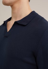 Polo din tricot bleumarin, cu o textură moale, având un guler ascuțit și o croială lejeră. Fără modele sau detalii metalice vizibile.