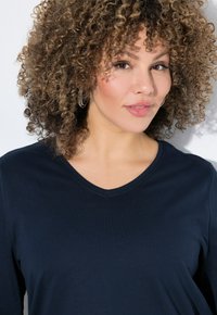 Marineblaue V-Ausschnitt-Shirt aus weichem Material mit langen Ärmeln und glatter Textur. Das Modell trägt dezente Ohrringe und hat natürliche Locken.