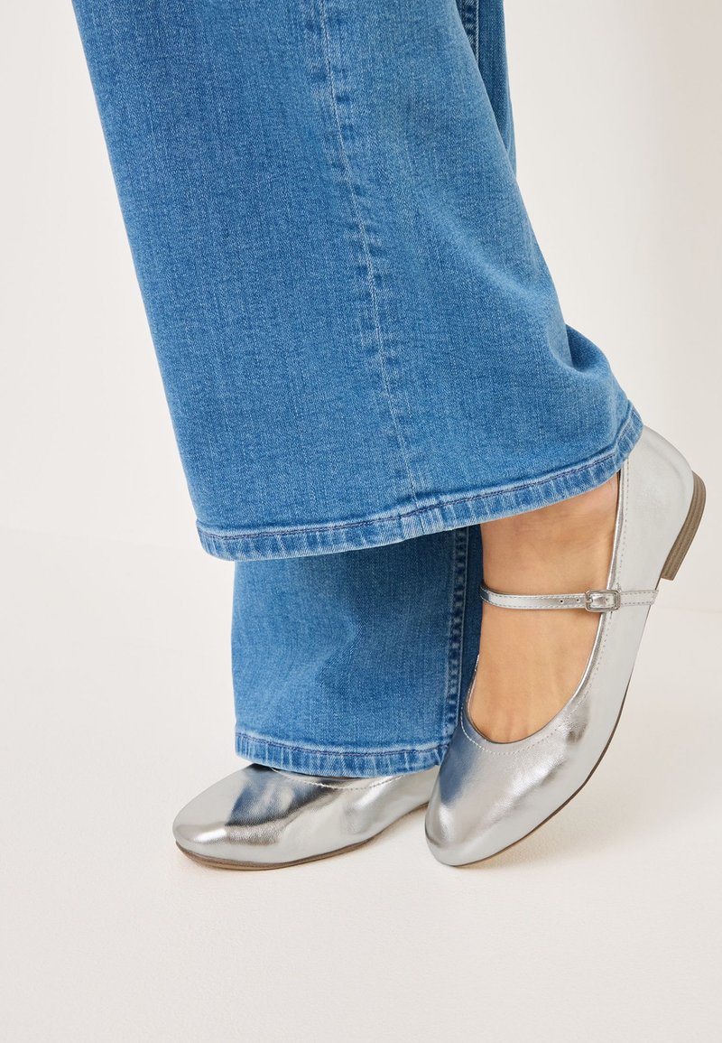 Sapatilhas de ballet metálicas prateadas com uma faixa no tornozelo, combinadas com calças de ganga largas azuis. Textura suave, ponta redonda, design minimalista.