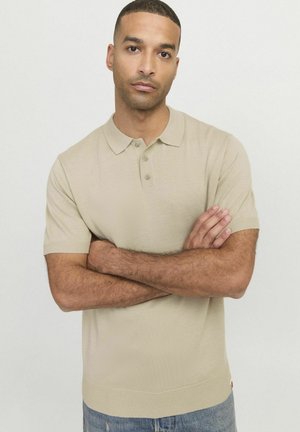 Polo beige à manches courtes avec col, patte de boutonnage à trois boutons et ourlet côtelé. Texture lisse avec une coupe ajustée. Jeans en denim visibles en dessous.