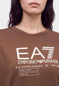 Γυναίκα που φοράει καφέ μπλουζάκι Emporio Armani EA7 με άσπρο κείμενο και λογότυπο, εμφανίζοντας τον λαιμό και το πάνω μέρος του στήθους.