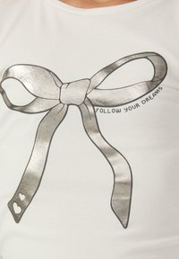 Witte T-shirt met een metallic zilveren strikafbeelding en de tekst "VOLG JE DROMEN." De strik heeft hartvormige accenten op het lint.