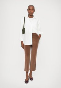 Pessoa confiante vestindo uma camisa branca de botão, calças curtas castanhas, sapatos bordô e carregando uma pequena bolsa verde à tiracolo.