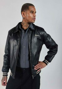 Veste en cuir noir avec fermeture éclair à l'avant, poignets et col côtelés. Présente une broderie "Redskins" sur la poitrine et des accents rayés sur les manches.