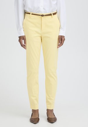 Isik kannab kitsalt istuvaid kahvatukollaseid pükse pruuni vööga, valget sisse toppitud särki ja pruune teravate ninadega madalaid jalanõusid, seistes lihtsa tausta ees.