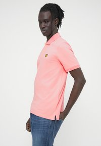 Ροζ μπλουζάκι πόλο με σταθερό γιακά, κοντά μανίκια και κίτρινο κεντημένο λογότυπο, συνδυασμένο με μπλε τζιν παντελόνι.