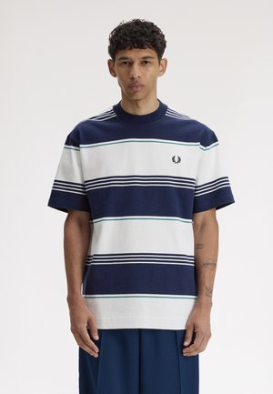 Jeune homme aux cheveux courts et bouclés portant un t-shirt à manches courtes rayé bleu marine et blanc avec un petit logo de laurier sur la poitrine.