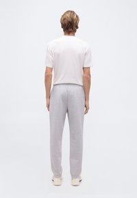 Άτομο με ανοιχτό καστανά μαλλιά στέκεται με την πλάτη προς τα εμπρός, φοράει λευκό κοντομάνικο πουκάμισο, ανοιχτογκρι φόρμα και λευκά παπούτσια σε απλό φόντο.