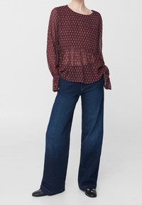 Blusa bordeaux a fantasia con maniche lunghe e silhouette a peplum, abbinata a jeans a gamba larga di colore blu scuro. Tessuto testurizzato e vestibilità rilassata.