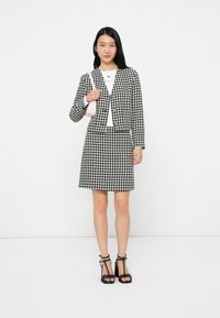 Fekete-fehér houndstooth öltöny, cropped zakóval és hozzá illő szoknyával. A modellen egy kis rózsaszín táska van, és fekete pántos magassarkút visel.