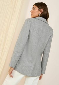 Šedý houndstooth blazer s přizpůsobeným střihem a klopovým límcem. Má dlouhé rukávy a texturovanou tkaninu, zobrazený zezadu.