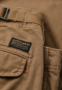 Nærbillede af brun stofbukselomme med sort og gul Superdry-mærkat og en syet bæltestrop.