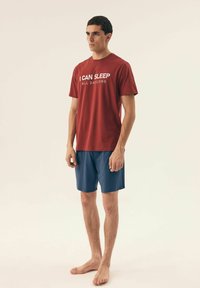 Czerwona bawełniana koszulka z białym napisem "MOGĘ SPAĆ CAŁY DZIEŃ", w połączeniu z niebieskimi krótkimi spodniami, stojąca na bosaka na neutralnym tle.