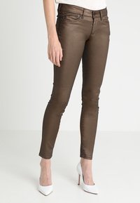 Vaqueros skinny metálicos marrones con una textura suave, que cuentan con un diseño clásico de cinco bolsillos y detalles de costura sutiles, llevados con tacones blancos.