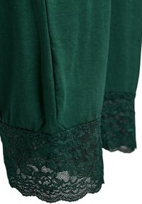 Temno zelena oblačila iz gladkega blaga z zapletenim čipkastim trakom na robu, ki prikazuje nežni vzorec in teksturirane detajle.