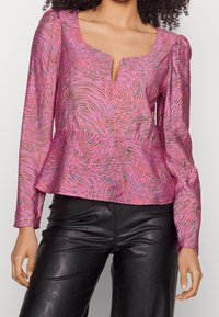 Donna che indossa una blusa a maniche lunghe rosa con un motivo e un collo a V piccolo, abbinata a pantaloni di pelle neri.