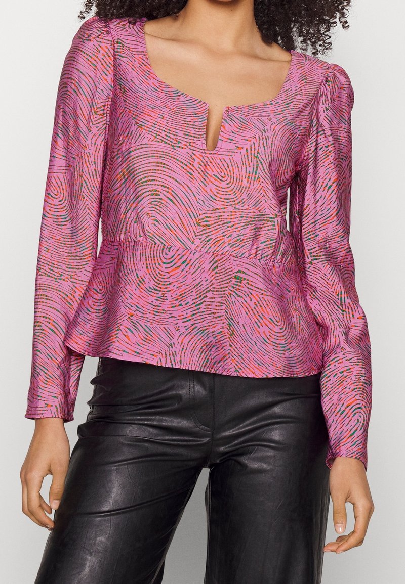 Donna che indossa una blusa a maniche lunghe rosa con un motivo e un collo a V piccolo, abbinata a pantaloni di pelle neri.