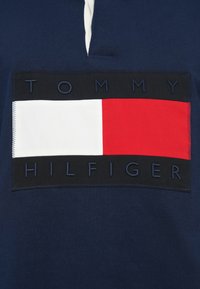 Μπλε ναυτικό πόλο πουκάμισο με μεγάλο έμβλημα λογότυπου σε κόκκινες, λευκές και μαύρες αποχρώσεις, με ανάγλυφο κείμενο που γράφει «TOMMY HILFIGER». Ύφασμα από βαμβάκι.