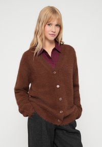 Cardigan castanho, fofinho, com decote em V e botões frontais. Textura suave, corte largo e mangas longas complementam calças escuras.