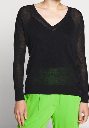 Pull noir à col en V à manches longues avec boutons latéraux, porté avec un pantalon taille haute vert vif, ne montrant que le torse et les bras.