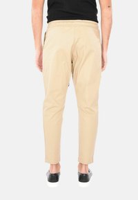 Pantaloni beige slim-fit con vita elastica, tasche laterali e una texture liscia. Sneakers nere con suole bianche sono indossate.