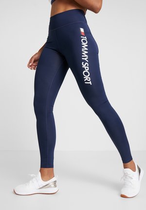 Laivastonsiniset leggingsit joustavasta kankaasta, joissa valkoinen teksti "TOMMY SPORT" sivussa. Yhdistetty valkoisiin urheilujalkineisiin.