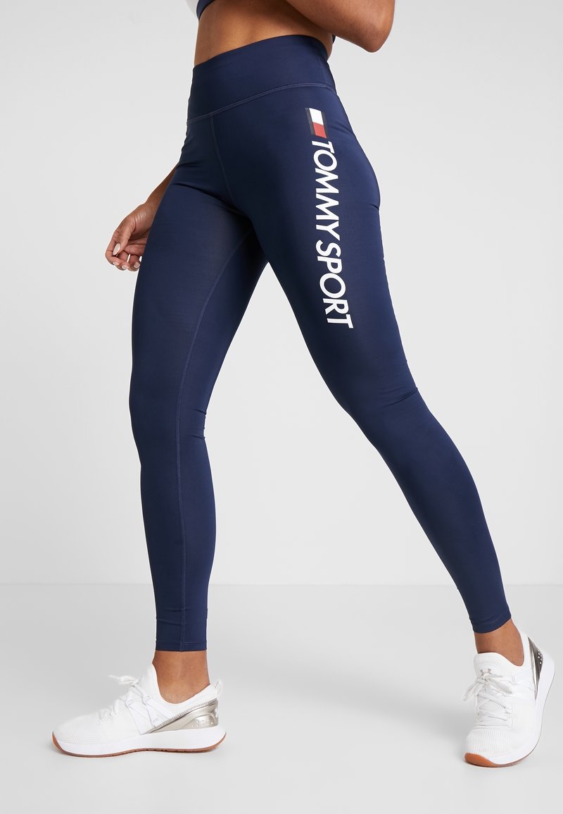 Granatowe legginsy wykonane z elastycznego materiału, z białym napisem "TOMMY SPORT" wzdłuż boku. Połączone z białymi butami sportowymi.