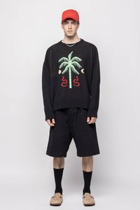 Svart strikkegenser med palmetrærdesign, røde detaljer, og oversized passform, kombinert med svarte shorts og rød caps. Lett strukturert stoff.