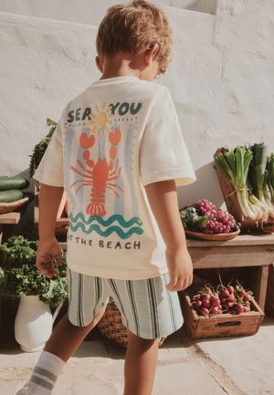 Barn iført en hvid skjorte med en hummer- og solmotiveret design og stribede shorts, står ved et markedbord med grøntsager og frugt.