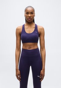 Αθλητική γυναίκα που στέκεται προς τα εμπρός, φοράει μοβ αθλητικό σουτιέν και ψηλόμεσα κολάν με λογότυπα Puma σε απλό φόντο.