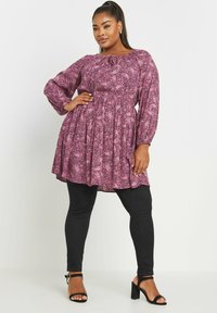 Tunika dengan pola paisley ungu, lengan panjang dan bodice smocked, dipadukan dengan legging hitam dan sandal hak hitam.