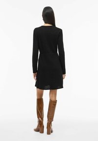 Robe noire en maille côtelée à manches longues, taille cintrée, ourlet évasé, présentée avec des bottes en cuir marron montant jusqu'au genou sur un fond uni.