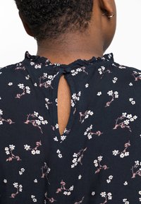 Mörkblå blus med blommönster och volang i halsringningen, med en nyckelhålsöppning på baksidan. Slät tyg med små vita och rosa blommor.