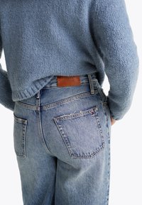 Svetlo modra pletena jopica v kombinaciji z visokim pasom modrimi jeans hlačami, ki imajo rjavo usnjeno označbo z vtisnjenim napisom "TOMMY HILFIGER".
