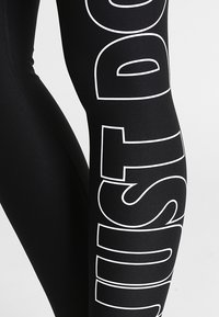 Leggings neri con testo bianco audace che recita "JUST DO IT" lungo il lato sinistro. Realizzati in un tessuto morbido e elasticizzato.