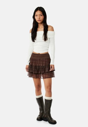 Vrouw draagt een witte off-shoulder top met lange mouwen, een bruine gerimpelde minirok, crèmekleurige sokken en donkerbruine kniehoge laarzen.