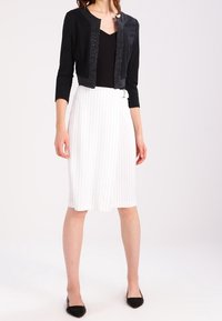 Czarna krótka kurtka ze srebrnym zdobieniem, zestawiona z białą spódnicą w cienkie paski. Strój uzupełniają czarne płaskie buty. Czyste linie i strukturalny design.