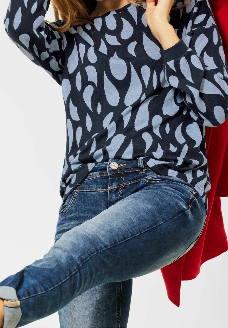 Personne portant une chemise à manches longues bleu à motifs et un jean bleu délavé levant une jambe, avec un vêtement rouge visible derrière eux.