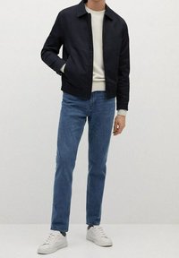 Eine Person trägt eine schwarze Zip-Jacke, einen cremefarbenen Pullover, blaue Jeans und weiße Sneaker und steht vor einem schlichten hellen Hintergrund.