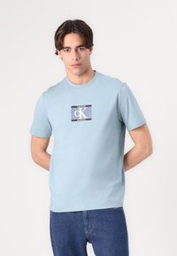 Lyseblå bomull t-skjorte med et grafisk merke som viser teksten "Calvin Klein" og "New York City" i kontrasterende farger. Korte ermer og rund hals.