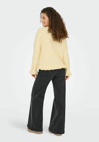 Pull jaune en tricot avec un ourlet et des manches volantés, associé à un jean noir taille haute à jambes larges, le tout sur un fond neutre.