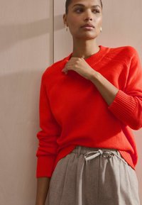 Punane kudumisko sweater v-kaelusega ja soonikkoes varrukaotstega, koos heledat hallidest, volditud pükstest, millel on kummist tõmbluku vöökoht.