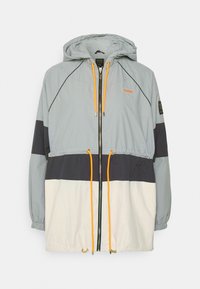 Chaqueta ligera con capucha, que presenta bloques de color en gris, azul marino y blanco. Incluye un cordón naranja, cierre de cremallera y puños elásticos.