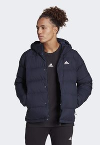 Marinblå puffjacka med huva, med sydda horisontella paneler, tryckknappar och vita Adidas-logotillbehör. Buren över en svart topp.