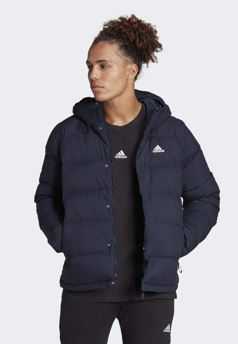 Marinblå puffjacka med huva, med sydda horisontella paneler, tryckknappar och vita Adidas-logotillbehör. Buren över en svart topp.