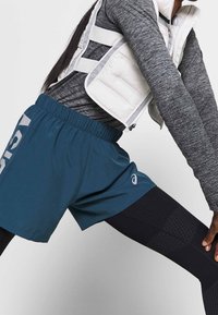 Athletic shorts in teal with a logo, paired with textured black tights and a grey long-sleeve top under a white vest.